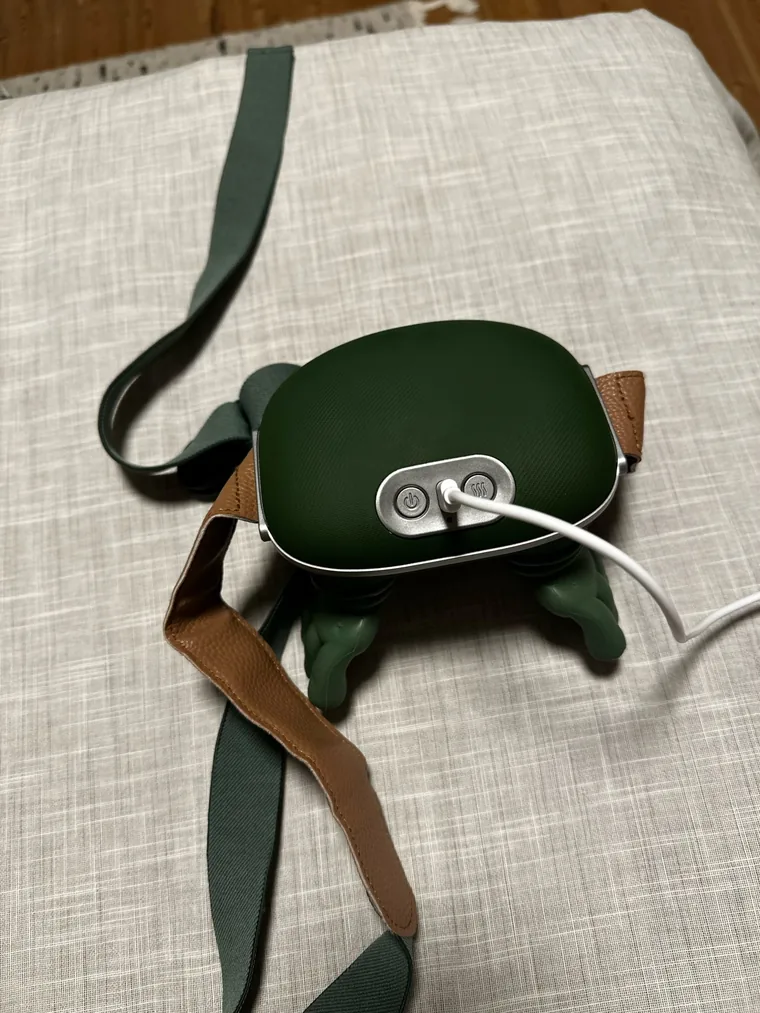 Le châle de massage Nuquea posé sur un coussin de lin, sa sangle verte et cuir déroulée, câble de charge branché